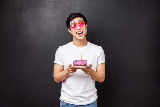Birthday, celebration and party concept. Enthusiastic cute asian guy celebrating b-day, tilt head and look happy camera with pleased smile, hold cake on plate with lit candle, making wish.