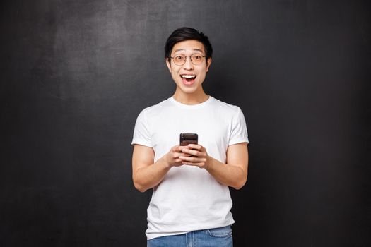 Technology, gadgets and people concept. Super happy smiling positive asian guy in glasses and t-shirt, react amused and wondered at awesome news received via email, hold mobile phone look camera.