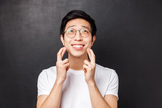 Close-up portrait of optimistic, hopeful young asian man cross fingers good luck, looking with hope and faith up sky, smiling praying to god, pleading dream come true, stand black background.