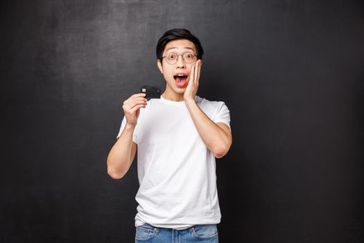 Bank, finance and payment concept. Portrait of excited and amused asian man in t-shirt, open mouth fascinated talking about credit card special features, standing black background intrigued.