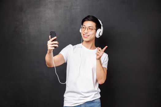 Technology, gadgets and people concept. Portrait of stylish young asian hipster guy in headphones, video-call friend using smartphone, taking selfie pointing finger right, black background.