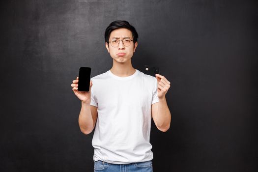 Bank, finance and payment concept. Portrait of miserable poor young asian male student showing his bank account empty on smartphone screen via application, hold credit card and complaining.