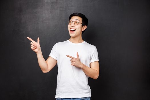 Portrait of enthusiastic and excited handsome chinese guy in white shirt, open mouth amused looking and pointing upper left corner over black background, spot great discounts.