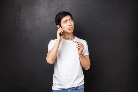 Technology, gadgets and people concept. Portrait of thoughtful and indecisive asian guy biting rim of glasses while thinking as talking on mobile phone, making hard choice what order over smartphone.