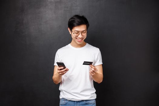 Bank, finance and payment concept. Portrait of cheerful asian man in t-shirt, holding mobile phone and credit card, insert billing info, buying products online, book tickets for flight with app.