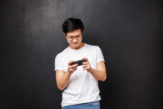 Technology, gadgets and people concept. Portrait of young asian guy hold mobile phone both hands horizontally, squinting and frowning as trying pass hard level in smartphone game.