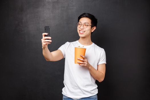 Portrait of handsome cheerful young asian male blogger record video of him eating popcorn and watching movie at cinema, taking selfie on smartphone smiling happy, black background.