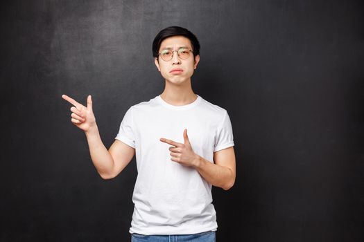 Portrait of skeptical and doubtful young serious smart guy thinking about proposal having doubts, pointing fingers left at advertisement and look camera suspicious with disbelief, black background.