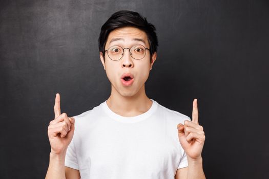 Close-up portrait of amused and excited happy asian man visit amusement park for first time, pointing fingers up at something super cool, say wow astonished, stand black background.