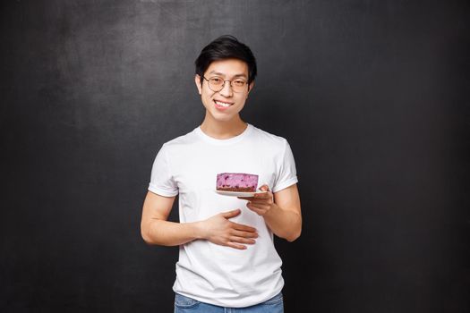 Portrait of pleased and staffed young cute asian man with piece of cake on plate, rubbing his belly and smiling satisfied, ate tasty dessert, delighted after having delicious meal, black background.