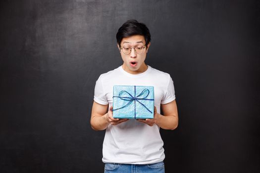 Celebration, holidays and lifestyle concept. Surprised and excited, amazed asian guy receive gift box, holding present and looking at it amused didnt expect coworker remembers about birthday.