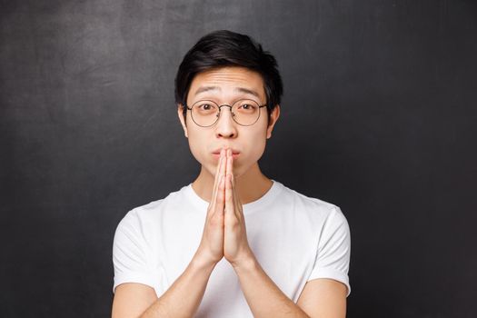 Close-up portrait of worried and uneasy hopeful young man in white t-shirt, glasses, hold hands in pray looking with pleading eyes, asking for help, begging forgiveness, black background.