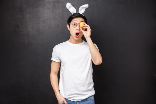 Holidays, party and Easter concept. Portrait of excited playful and funny asian young man in rabbit ears, holding painted egg over eye and looking amused with opened mouth at camera.