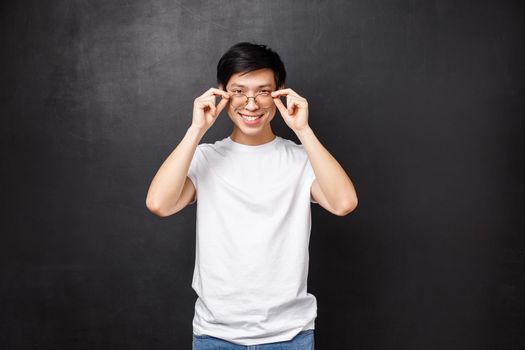 Sassy handsome young asian guy picking new prescribed glasses in optician store, put on eyewear and look confident and pleased camera, smiling white teeth, black background.