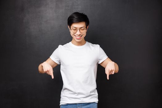 Portrait of cheerful friendly-looking asian hipster guy in sunglasses and white t-shirt spot cool promo discount, got best deal, pointing and looking down satisfied, stand black background.