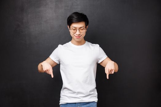 Portrait of satisfied and happy smiling young male student, asian guy in white t-shirt looking and pointing down, indicate bottom advertisement, found something cool, black background.