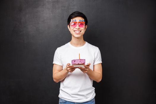 Birthday, celebration and party concept. Happy young cheerful asian guy in white t-shirt, funny glasses holding b-day cake with candle, blow it to make a wish, smiling excited, stand black background.