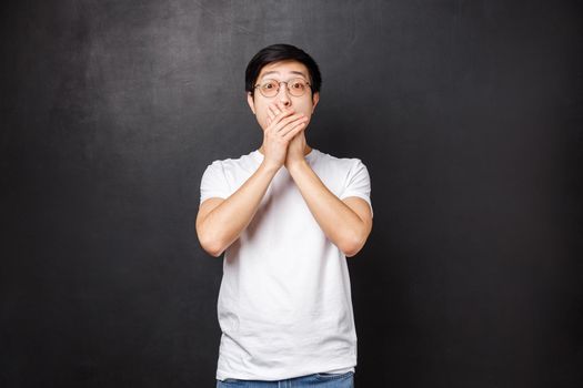 Lifestyle and people concept. Speechless alarmed and scared young asian guy found out something scary, gasping cover mouth in panic, staring camera popped eyes full of fear, black background.