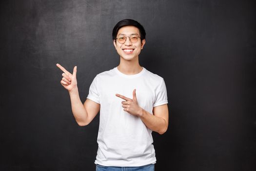 Portrait of good-looking young asian man, male student in white shirt pointing fingers upper left corner at promo, discount offer or banner, smiling camera, suggest visit link, black background.
