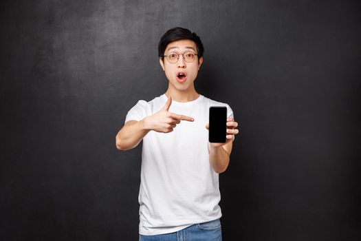 Technology, gadgets and people concept. Impressed and excited handsome asian guy telling to download app, check and see awesome application in your mobile store, point at smartphone screen.