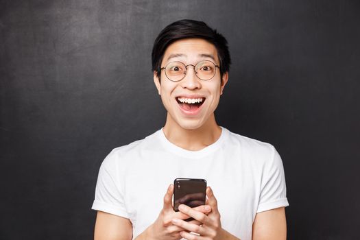 Technology, messaging and people concept. Close-up portrait of excited and amused happy asian man, smiling astonished and pleased, received positive reply in mobile phone messanger, hold mobile.