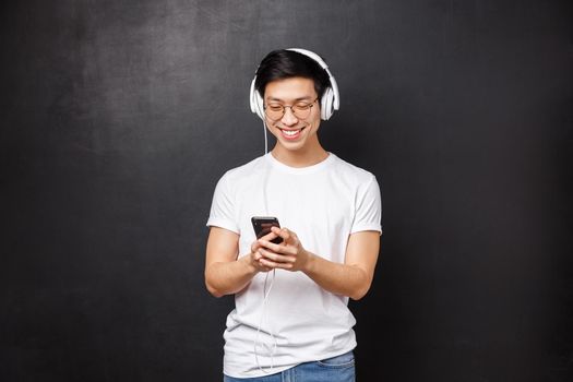 Technology, gadgets and people concept. Handsome happy young smiling asian man in t-shirt, listen music in headphones, pick playlist in mobile phone, texting friend, black background.