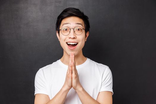 Close-up portrait of hopeful and grateful young happy asian guy receive help after begging friend, saying thank you, hold hands in pray, appreciate gesture, standing black background.