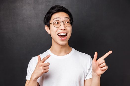 Close-up portrait of excited and happy, cheerful asian guy looking at something incredibly awesome, pointing and staring right side, smiling astounded and fascinated, black background.