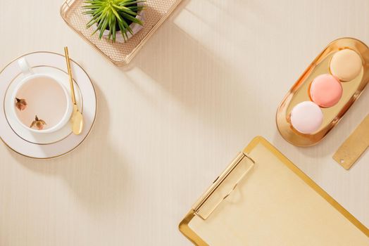 Modern gold stylized home office desk with folder, macaroons, coffee mug on beige background. Flat lay, top view lifestyle concept.`