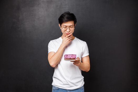 Portrait of thoughtful and doubtful asian guy looking hesitant at piece of cake, thinking eat or leave it in the fridge, standing over black background, being on diet, allergic on ingridients.