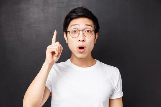 Close-up portrait of smart and creative asian man in glasses, raising index finger add suggestion, saying his opinion or thought, want to ask question, invented good plan, black background.