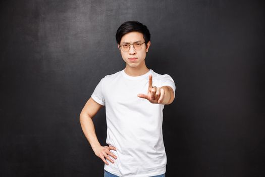 Portrait of serious-looking and displeased young asian guy give warning or caution sign, extend and shake finger as scolding forbid action, dont like idea and prohibit do it, black background.