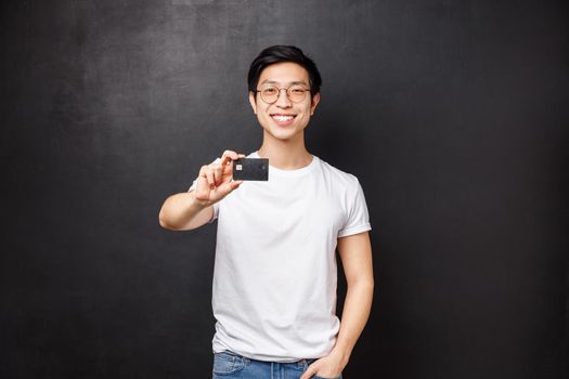 Bank, finance and payment concept. Portrait of friendly confident young asian male showing credit card at camera with pleased beaming smile, advice open account at put money, black background.
