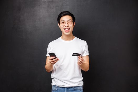 Bank, finance and payment concept. Portrait of amused handsome young asian guy holding credit card and mobile phone, smiling at camera, like shopping online, open new account, black background.