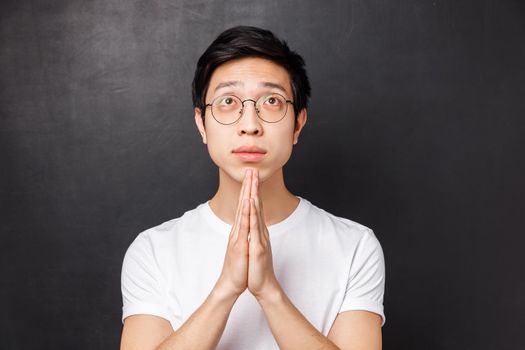 Close-up portrait of hopeful dreamy young asian guy in glasses and white t-shirt, hold hands together in pray, looking up pleading, anticipating miracle, awaiting results with hope.