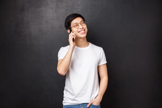 Technology, gadgets and people concept. Carefree cheerful asian man talking on phone with friendly attitude, hold hand in pocket casual pose, smile and look away, hold smartphone near ear.