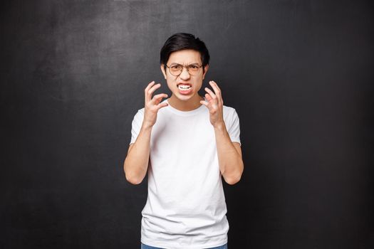 Aggressive young asian guy looking with hate and disgrace, cursing someone, feel angry and rage, clench hands furious want kill or punch someone in face, grimacing, black background.