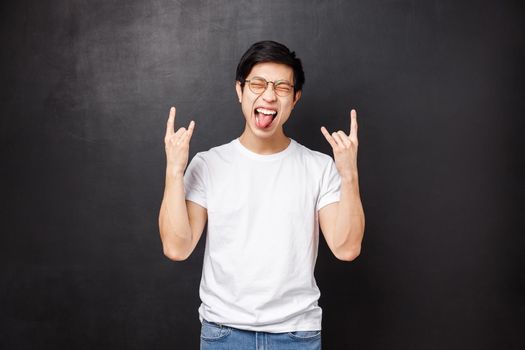 Happy excited and outgoing asian guy in glasses and t-shirt, having fun show tongue close eyes relaxed and make rock-n-roll heavy metal gesture as listening to awesome song, black background.