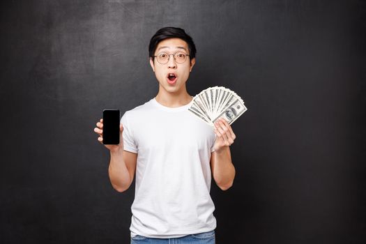 Technology, money and prizes concept. Amazed and surprised asian guy holding dollars, big cash and mobile phone, showing screen of smartphone banking application, look shocked camera.