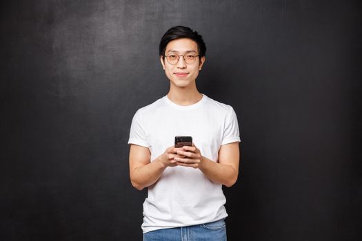 Technology, gadgets and people concept. Handsome young determined asian guy in t-shirt and glasses using mobile phone application, hold smartphone and smile at camera, black background.