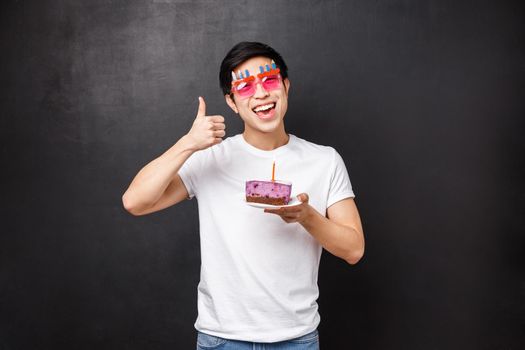 Birthday, celebration and party concept. Excited funny asian man in silly glasses enjoying b-day holding cake with candle, making wish laughing and smiling, show thumbs-up, black background.