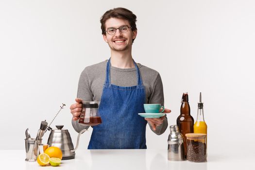 Barista, cafe worker and bartender concept. Portrait of friendly pleasant smiling man in apron give customer cup of brewed filter coffee, holding drink and kettle, white background.