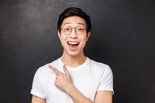 Close-up portrait of enthusiastic asian male coworker having lively conversation, discuss event or new product in stock, pointing finger upper left corner or behind, black background.