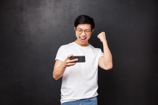 Technology, gadgets and people concept. Happy joyful lucky asian guy fist pump in celebration, hold mobile phone and smiling say yes, triumphing passed game or level, black background.