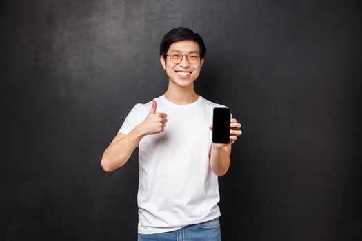 Technology, gadgets and people concept. Assertive cute asian guy in t-shirt and glasses, recommend online store or application for mobile phone, show thumbs-up and smartphone display.