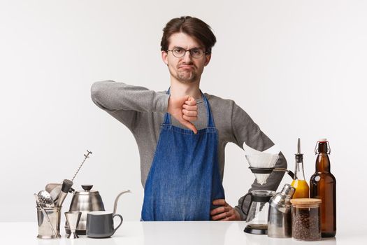 Barista, cafe worker and bartender concept. Skeptical and displeased bearded man employee in apron, smirk disappointed and show thumb-down in dislike, standing near coffee bar counter.