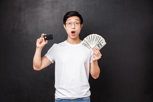 Technology, money and prizes concept. Excited and amazed rich lucky asian guy winning money, receive reward, holding dollars and credit card with overwhelmed astonished expression.