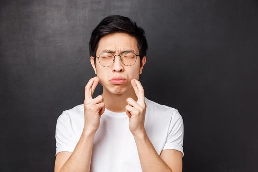 Close-up portrait of embarrassed and cautious, worried asian man in white t-shirt, close eyes pouting nervous while cross fingers good luck, praying, want dream come true, begging faith.