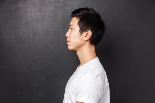 Profile portrait of young asian male model in white t-shirt smiling casually standing and looking left over black background of studio, waiting in queue, concept of people and lifestyle.