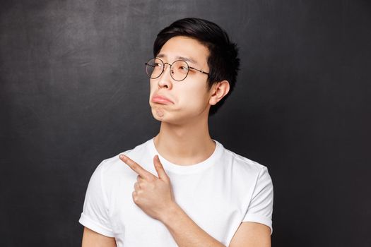 Close-up portrait of jealous and gloomy asian young man feel regret, look at something upsetting, pouting pointing finger left, desire have something but cant afford it, black background.